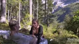 Frau mit langen blonden Haaren kniet im Schnee und bläst einen Mann vor Bergkulisse