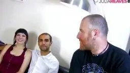 Junges spanisches Paar sitzt eng zusammen auf schwarzer Ledercouch, sie im roten Top, er im weißen Hemd, beide lächeln verlegen in die Kamera