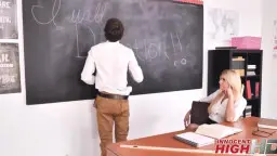 Blonde Lehrerin steht streng vor dem Pult, während ihr Schüler sich von hinten an sie drückt