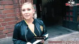 Blonde Lederjacken-Blondine mit tiefem Ausschnitt steht vor roter Backsteinmauer und lächelt verführerisch in die Kamera